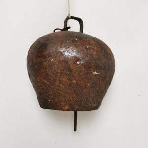 May include: A rustic, brown metal bell with a weathered appearance. The bell is suspended by a metal loop and a thin string. The bell has a rounded shape with a clapper hanging from the bottom. The bell is likely used for livestock or decorative purposes.
