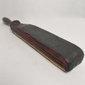 May include: A vintage leather strop with a wooden handle. The strop is black with red and yellow stitching. It is used for sharpening straight razors.
