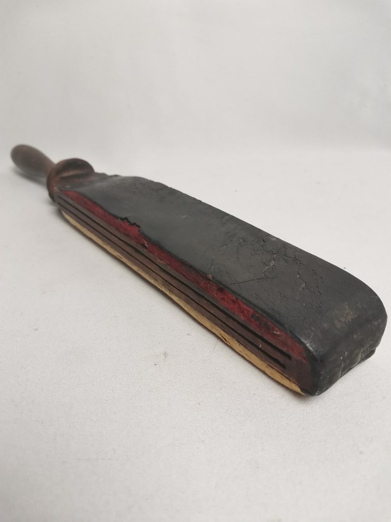 May include: A vintage leather strop with a wooden handle. The strop is black with red and yellow stitching. It is used for sharpening straight razors.