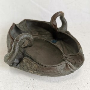 May include: A dark brown, antique-style ashtray with a unique, organic shape. The ashtray features a central bowl with raised edges and decorative, curved handles. The surface has a textured, weathered appearance, suggesting age and use.