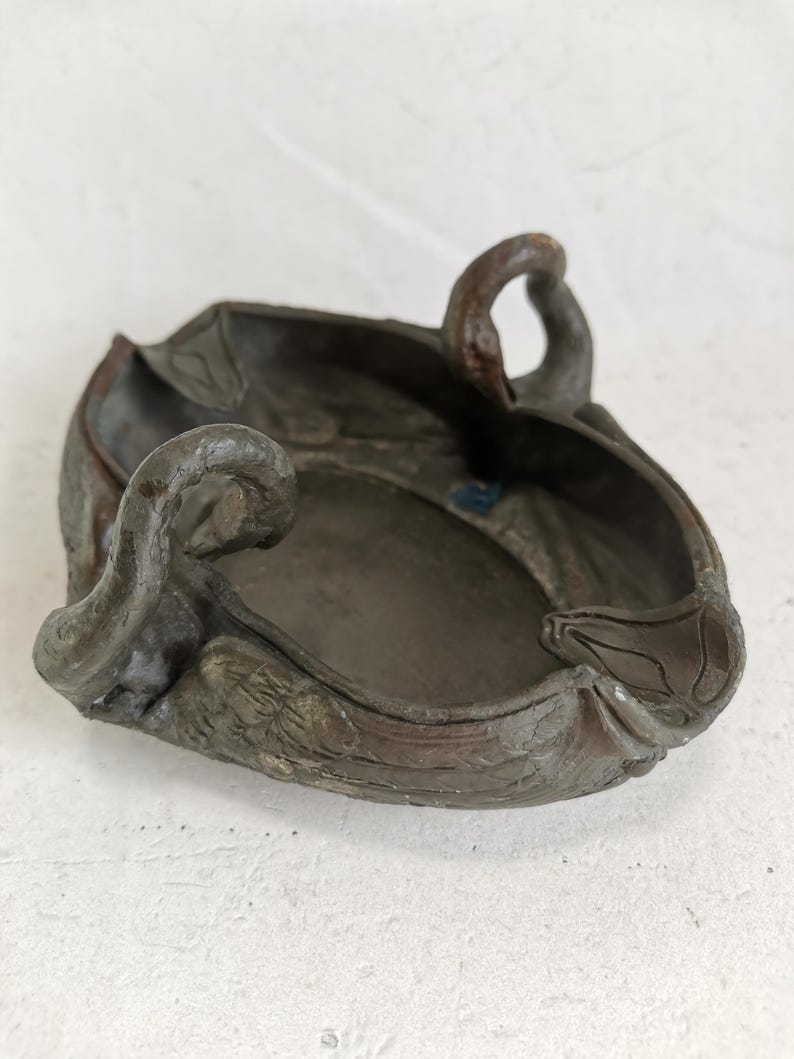 May include: A dark brown, antique-style ashtray with a unique, organic shape. The ashtray features a central bowl with raised edges and decorative, curved handles. The surface has a textured, weathered appearance, suggesting age and use.