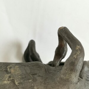 May include: A weathered, dark bronze sculpture of a reclining figure with outstretched arms. The figure is resting in a shallow, rectangular basin. The sculpture has a textured surface and appears to be made of metal.