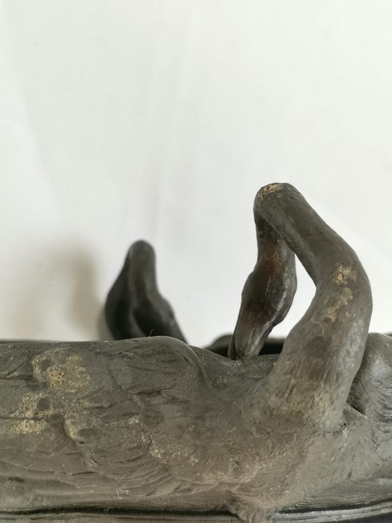 May include: A weathered, dark bronze sculpture of a reclining figure with outstretched arms. The figure is resting in a shallow, rectangular basin. The sculpture has a textured surface and appears to be made of metal.
