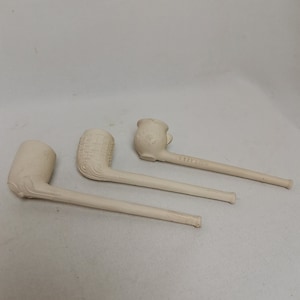 May include: Three off-white ceramic tobacco pipes. The pipes have different bowl designs, including a textured pattern and floral details. Each pipe has a long, slender stem.
