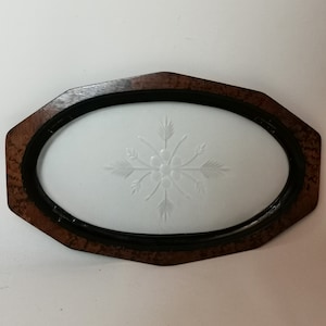 Puede incluir: Una bandeja ovalada de madera con un borde negro y un inserto de vidrio esmerilado blanco. El inserto de vidrio tiene un diseño de copo de nieve grabado.