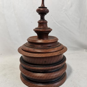 May include: A dark brown, turned wooden box with a lid. The box is made of stacked, rounded sections, and has a decorative finial on top. The wood grain is visible, and the piece has a classic, antique aesthetic.