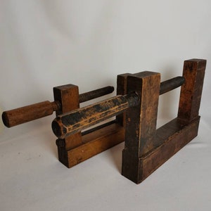 May include: A vintage wooden clamp with a threaded screw and a wooden handle. The clamp is made of dark wood and has a rustic appearance.