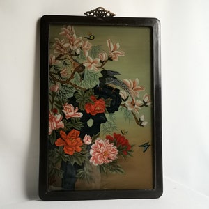Può includere: Un dipinto incorniciato che raffigura un'arrangiamento floreale con fiori di magnolia rosa e bianchi, peonie rosse e arancioni e foglie verdi. Due uccelli sono appollaiati su un ramo e farfalle volano intorno ai fiori. La cornice è nera con un accento dorato.