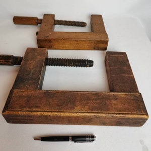 May include: Two vintage wooden C-clamps with a black metal screw. The clamps are made of dark brown wood and have a worn, rustic look.