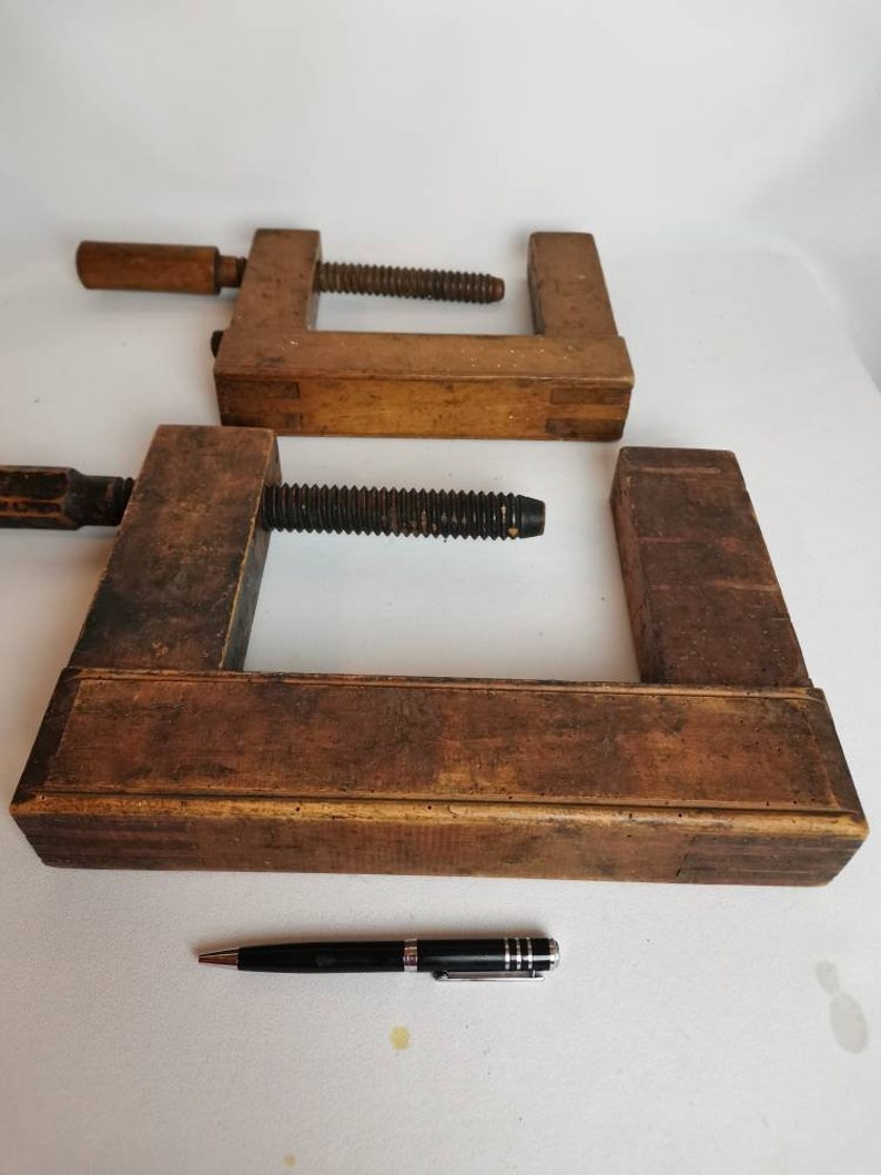 May include: Two vintage wooden C-clamps with a black metal screw. The clamps are made of dark brown wood and have a worn, rustic look.