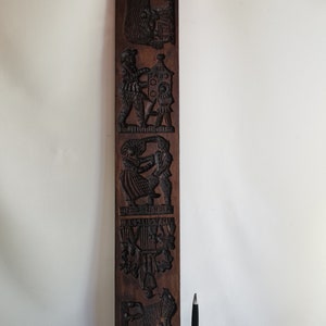 May include: A vintage wooden cookie mould with intricate carvings of various scenes, including a bear, a man with a lantern, a couple dancing, a group of people, and a horse. The mould is brown and has a rustic appearance.