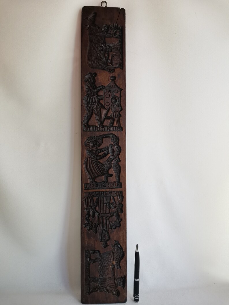 May include: A vintage wooden cookie mould with intricate carvings of various scenes, including a bear, a man with a lantern, a couple dancing, a group of people, and a horse. The mould is brown and has a rustic appearance.