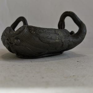 May include: A dark gray, swan-shaped bowl with a textured surface. The bowl has two swan heads forming handles and a detailed body. The bowl is likely made of metal or a similar material.