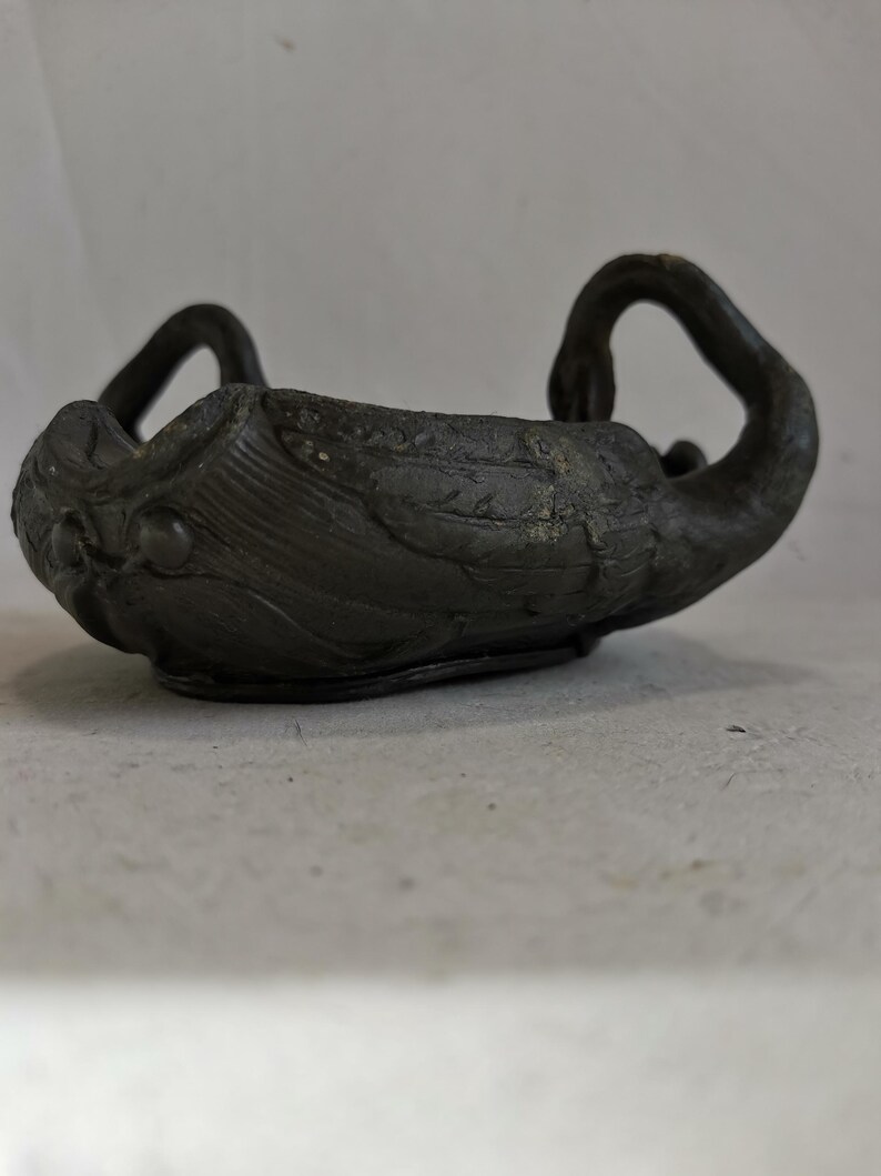 May include: A dark gray, swan-shaped bowl with a textured surface. The bowl has two swan heads forming handles and a detailed body. The bowl is likely made of metal or a similar material.