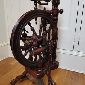 May include: Antique wooden spinning wheel with a dark brown finish. The wheel has a large wooden wheel with spokes and a smaller wheel with a spindle. The spinning wheel is on a wooden base with three legs.