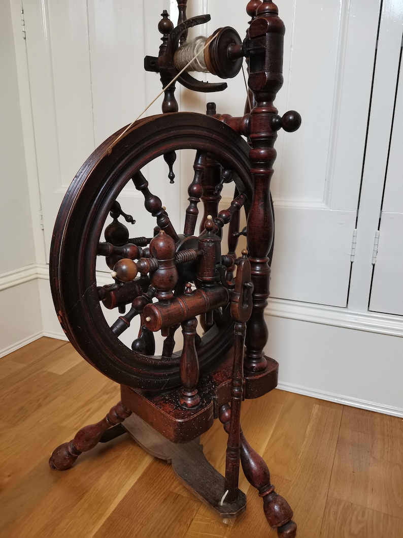 May include: Antique wooden spinning wheel with a dark brown finish. The wheel has a large wooden wheel with spokes and a smaller wheel with a spindle. The spinning wheel is on a wooden base with three legs.