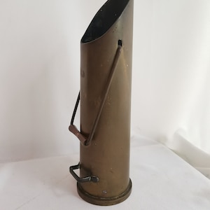 Pode incluir: Um porta-lenha de lareira de latão com uma alça de metal preto. O porta-lenha é alto e cilíndrico com uma parte superior inclinada.