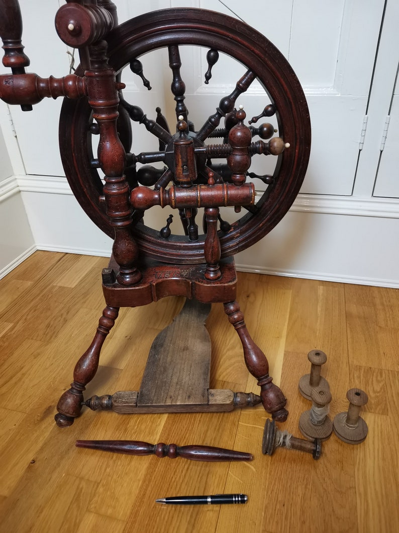 May include: A wooden spinning wheel with a large, dark brown wheel and a wooden base. The wheel has a intricate design with spokes and a central hub. There are several wooden spools and a pen on the floor next to the wheel.