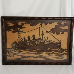 May include: A framed wooden artwork depicting a vintage cruise ship sailing on wavy waters. The ship is dark brown, with detailed cut-out windows and smokestacks. The background is a lighter wood, with dark brown clouds. The frame is a dark brown.