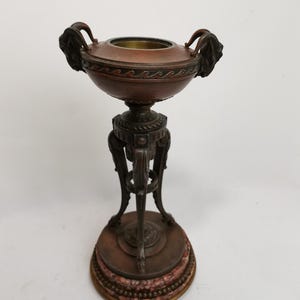 May include: A bronze decorative stand with a round top and three legs. The stand has a marble base and features a decorative design.