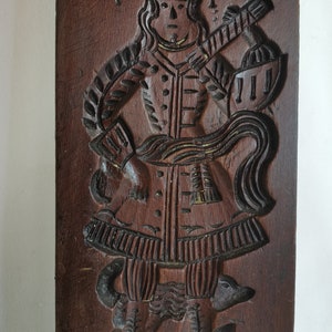 May include: A wooden cookie mould with a detailed carving of a person in a long coat and hat standing on two animals. The mould is brown and has a worn, antique look.