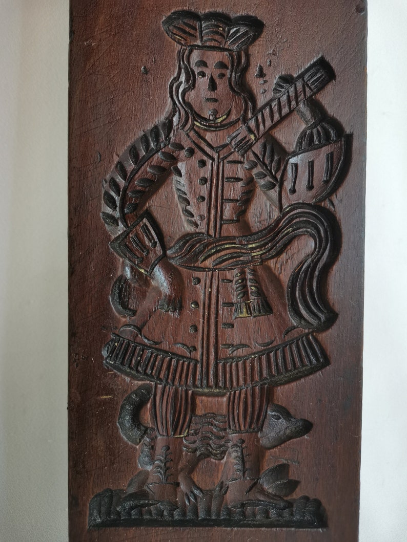 May include: A wooden cookie mould with a detailed carving of a person in a long coat and hat standing on two animals. The mould is brown and has a worn, antique look.