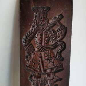 May include: A vintage wooden cookie mould featuring a detailed carving of a man in a hat and coat, standing with a dog at his feet.