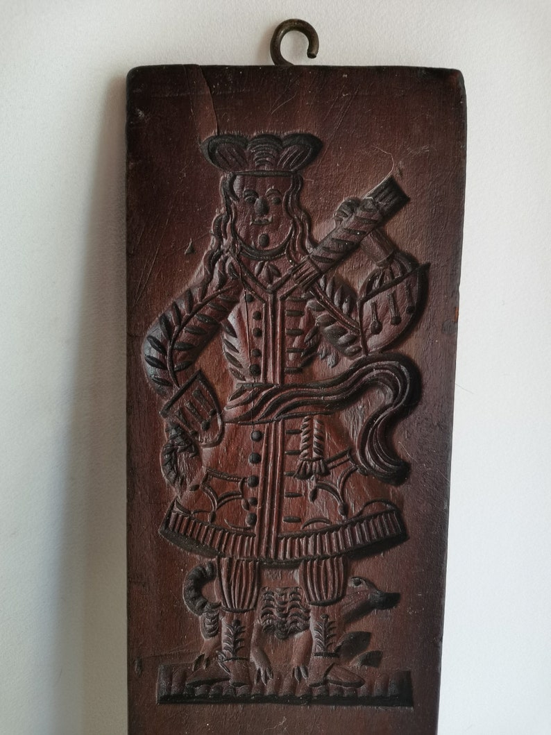 May include: A vintage wooden cookie mould featuring a detailed carving of a man in a hat and coat, standing with a dog at his feet.