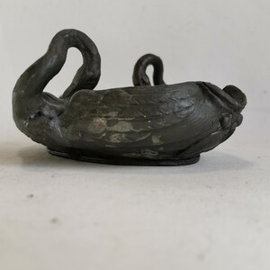 May include: A dark, antique-style swan-shaped bowl. The bowl is made of a dark metal with a textured surface. The swan's neck curves upward, and the bowl has a rounded base. The overall design is detailed and elegant.