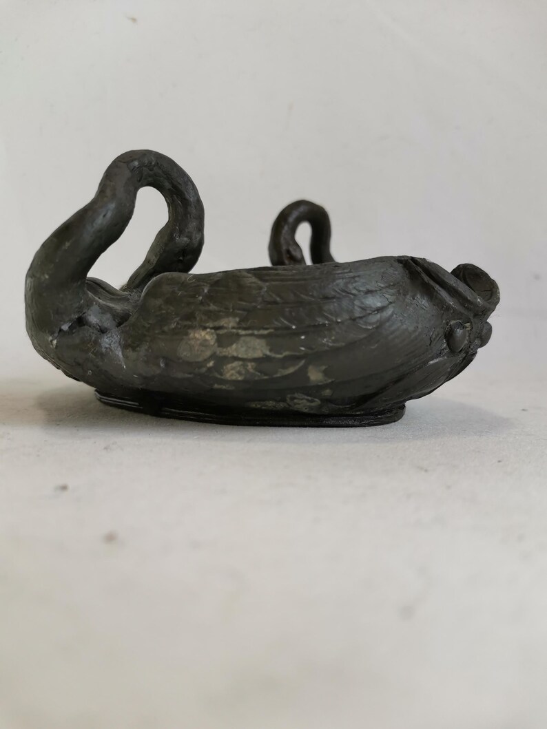 May include: A dark, antique-style swan-shaped bowl. The bowl is made of a dark metal with a textured surface. The swan's neck curves upward, and the bowl has a rounded base. The overall design is detailed and elegant.