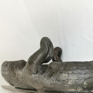 May include: A dark gray metal swan-shaped wine bottle holder. The swan's neck curves upward, and its wings are detailed. The holder is designed to cradle a wine bottle, adding a decorative touch to a table setting.