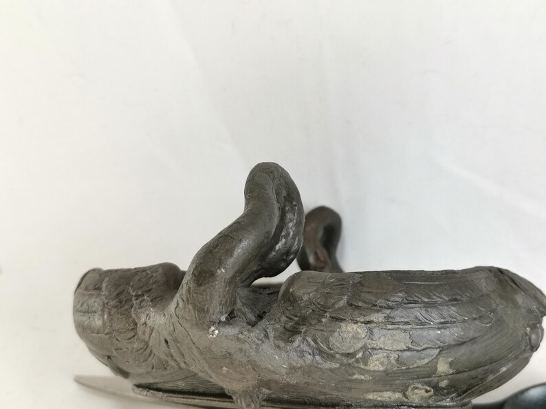 May include: A dark gray metal swan-shaped wine bottle holder. The swan's neck curves upward, and its wings are detailed. The holder is designed to cradle a wine bottle, adding a decorative touch to a table setting.