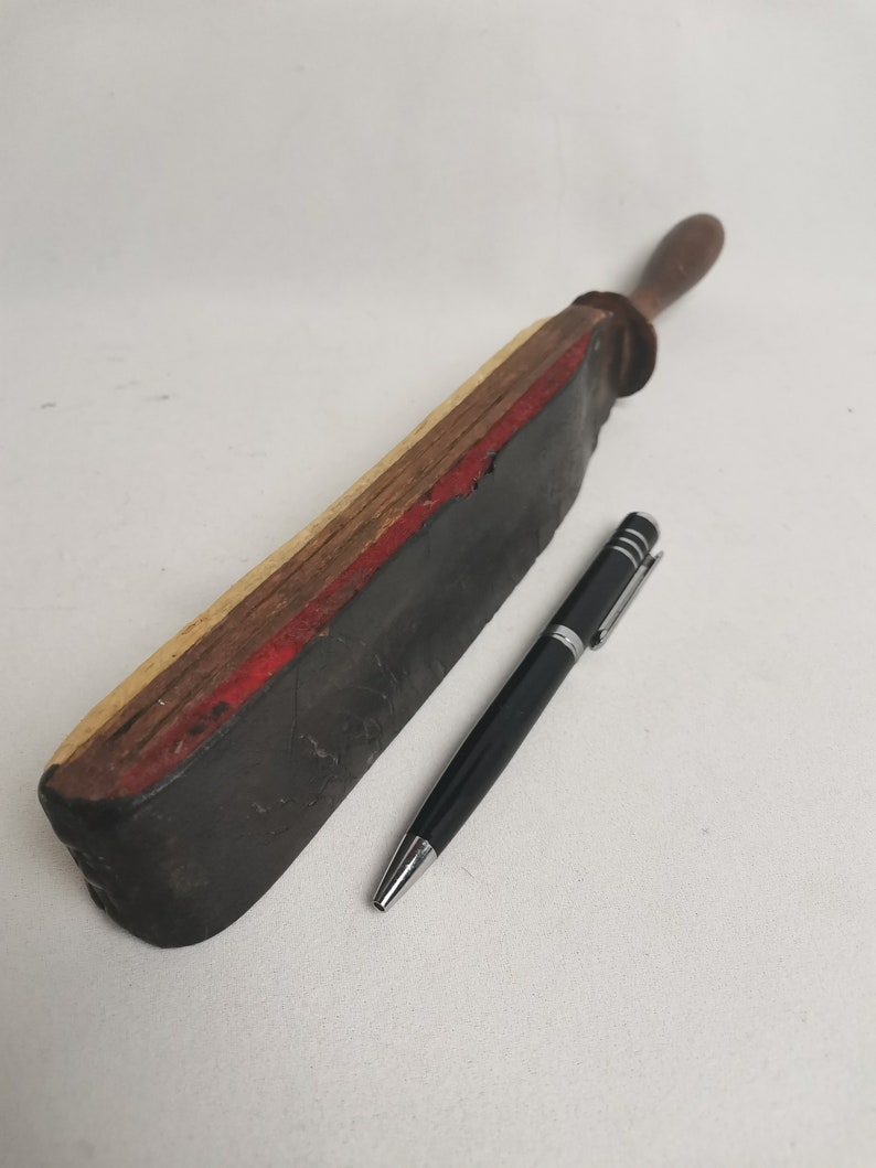 May include: A vintage leather strop with a wooden handle. The strop is made of layered wood and has a red stripe. The strop is about 12 inches long and 2 inches wide.