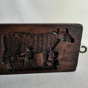 May include: A vintage wooden cookie mould with a carved design of a cow being milked by a person. The mould has a metal hook on the side.