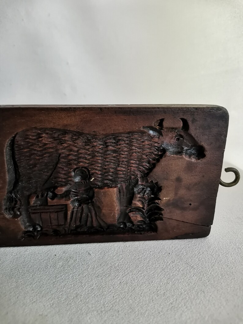 May include: A vintage wooden cookie mould with a carved design of a cow being milked by a person. The mould has a metal hook on the side.