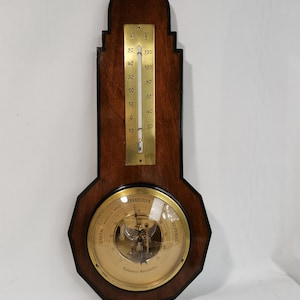 May include: A vintage wooden weather station featuring a thermometer and barometer. The thermometer, in Fahrenheit, is positioned above the barometer. The barometer has a glass cover and a brass face. The measurements are in Fahrenheit.