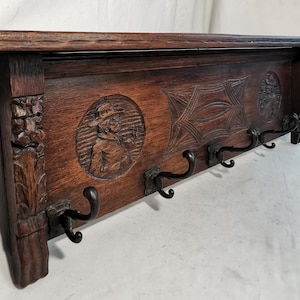 Peut inclure: Un porte-manteau en bois brun foncé avec des détails sculptés et cinq crochets en métal noir. Le porte-manteau comporte une étagère et des sculptures décoratives, dont un relief circulaire d'une personne portant un chapeau. Le bois a un aspect riche et vieilli.