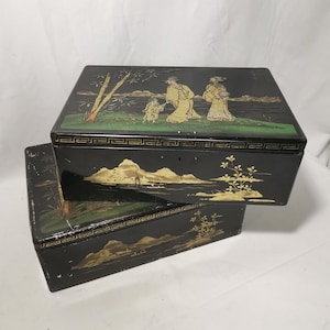 Peut inclure: Deux boîtes décoratives noires avec des scènes de paysage peintes en or et vert. La boîte supérieure présente des personnages en robes jaunes, du bambou et une scène de lac. La boîte inférieure a un design de paysage similaire. Les deux boîtes sont rectangulaires.
