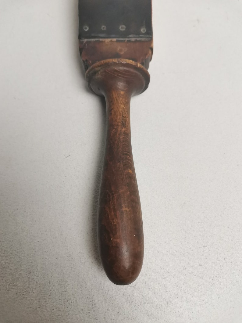 May include: A wooden strop with a dark brown leather surface and a wooden handle. The leather surface has a row of small metal rivets.