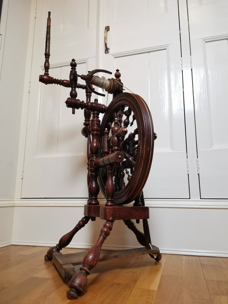 May include: A wooden spinning wheel with a dark brown finish. The wheel has a large, round wooden frame with a smaller, round wheel in the center. The spinning wheel is standing on a wooden base with four legs.