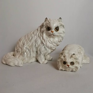 Puede incluir: Dos figuritas de porcelana blanca de gatos persas. El gato más grande está sentado con la cola enroscada alrededor de su cuerpo. El gato más pequeño está acostado con la cabeza levantada. Ambos gatos tienen ojos dorados y están pintados con un patrón de pelaje blanco y gris.