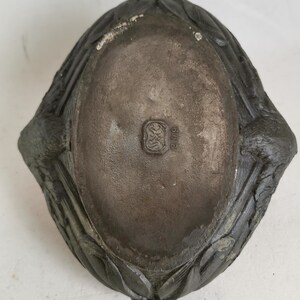 May include: A dark gray, oval-shaped dish with a textured surface. The dish has a decorative, leaf-like border and a small, embossed emblem in the center. The dish appears to be made of metal.