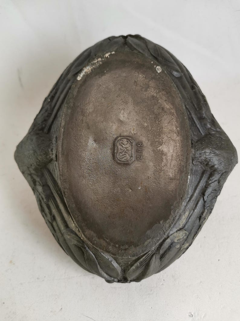 May include: A dark gray, oval-shaped dish with a textured surface. The dish has a decorative, leaf-like border and a small, embossed emblem in the center. The dish appears to be made of metal.