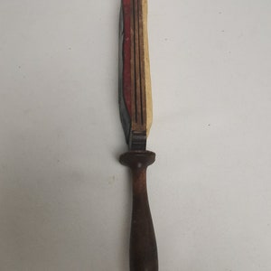 May include: A wooden tool with a long, narrow handle and a rectangular head with multiple layers of red, white, and black paint. The tool is likely used for sharpening or smoothing.