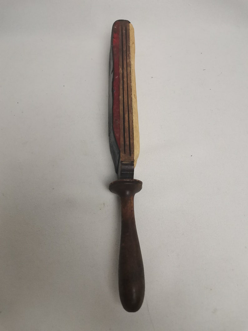 May include: A wooden tool with a long, narrow handle and a rectangular head with multiple layers of red, white, and black paint. The tool is likely used for sharpening or smoothing.