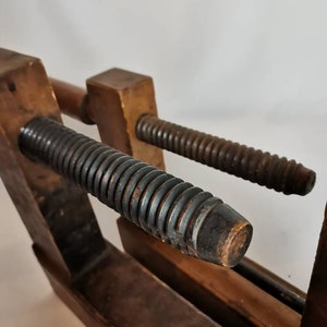 May include: A vintage wooden clamp with two threaded metal screws. The clamp is made of dark wood and has a rustic appearance.