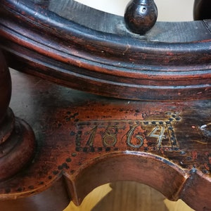 May include: Antique wooden spinning wheel with a dark brown finish and a red stain. The wheel has a date of "1864" inscribed on the base.
