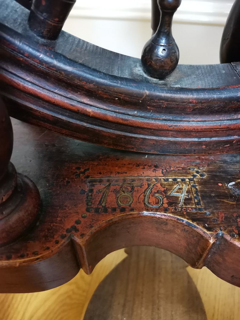 May include: Antique wooden spinning wheel with a dark brown finish and a red stain. The wheel has a date of "1864" inscribed on the base.