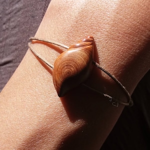 May include: A wooden seashell charm bracelet with a brown cord. The charm is carved from wood and has a natural wood grain pattern. The bracelet is tied around a person's wrist.