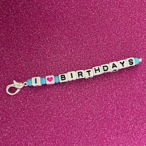 May include: A white beaded keychain with the words "I love birthdays" spelled out in black letters. A pink heart bead is between the "I" and "love" beads.